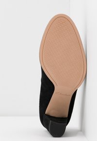 Svart mockaskor med spetsig tå och låg klack. Sulan är ljusbrun med en strukturerad yta. Logotypen "Clarks" är synlig på sulan.