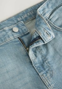 Pantaloni din denim slim fit, de culoare albastră deschisă, cu un nasture argintiu, închidere cu fermoar și textură decolorată; prezintă detalii de cusătură și o etichetă în interior.