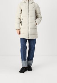 Puffet lysegrå frakke med hætte, struktureret fløjlstof, lynlås lommer og lige snit, parret med blå jeans og sorte sko.