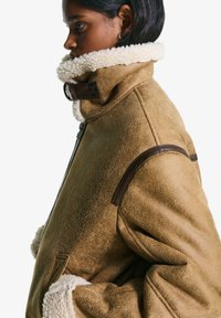 Brun shearlingjacka med krämfärgad pälskrage och kant, dragkedja fram och mörkbruna läderdetaljer på axlarna.