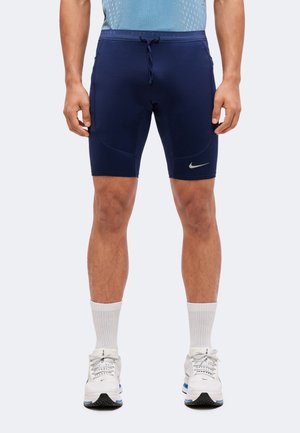 Homme portant un short de sport bleu marine avec le logo Nike, des chaussettes blanches hauteur cheville et des chaussures de course blanches, debout les bras relâchés le long du corps.
