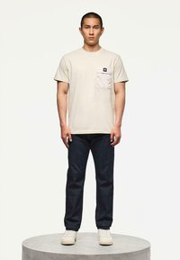 Mężczyzna stojący na okrągłym piedestale, ubrany w beżowy T-shirt z krótkim rękawem i kieszonką na piersi, ciemne dżinsy i białe trampki, na jednolitym tle.