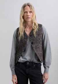 Chaleco de mezcla de lana en patrón de espiga marrón con botones oscuros, usado sobre una camisa de manga larga gris y combinado con jeans negros.