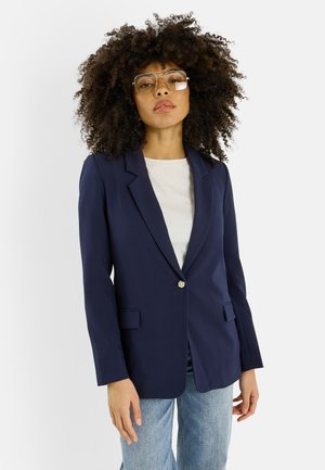 Femme aux cheveux bouclés et volumineux portant de grandes lunettes carrées, un blazer bleu marine à un bouton, une chemise blanche et un jean bleu clair.