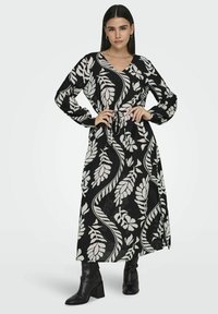 Musta maxi-mekko valkoisella lehväkuviolla, V-pääntiellä, pitkät hihat, kiristetty vyötärö ja sivuaukot, yhdistettynä polven yli ulottuviin mustiin korkokenkiin.