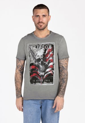 Uomo con capelli corti e tatuaggi indossa una t-shirt grigia con una grafica di teschio e bandiera americana con la scritta "La libertà non è mai gratis."