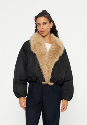 Femme portant une veste noire avec un grand col en fausse fourrure beige et un pantalon foncé, debout sur un fond blanc uni.
