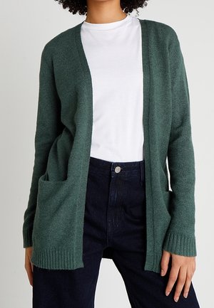 Femme portant un cardigan vert foncé ouvert sur le devant avec des poches, un t-shirt blanc et un jean bleu foncé sur un fond uni.