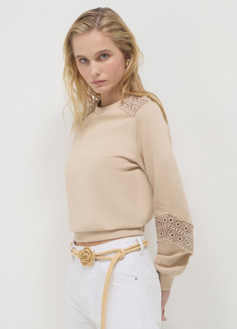 Felpa beige con dettagli in pizzo sulle spalle e maniche. Presenta polsini a coste e una vestibilità rilassata. Abbinata a pantaloni bianchi e una cintura annodata.