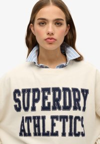 Sudadera cremosa con el texto en navy "SUPERDRY ATHLETICS", superpuesta a una camisa de cuello a rayas azul claro. Tejido suave; ajuste relajado.
