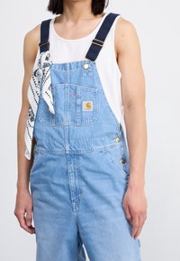 Hellblaue Jeanslatzhosen mit dunkelblauen Trägern, goldener Hardware und einem Logo-Patch. Ein weißes Bandana ist am linken Träger gebunden.