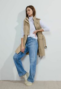 Beige ärmellose Weste mit Druckknöpfen, kombiniert mit einem weißen Rollkragenpullover und hellblauen Jeans. Das Model trägt beige Sneakers.