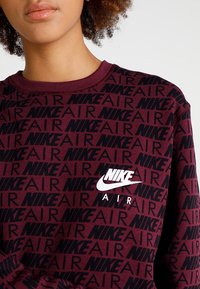 Burgundy sweatshirt med svart upprepande "NIKE AIR"-text, med en vit Nike-logotyp på bröstet. Mjuk tyg och ribbad krage.