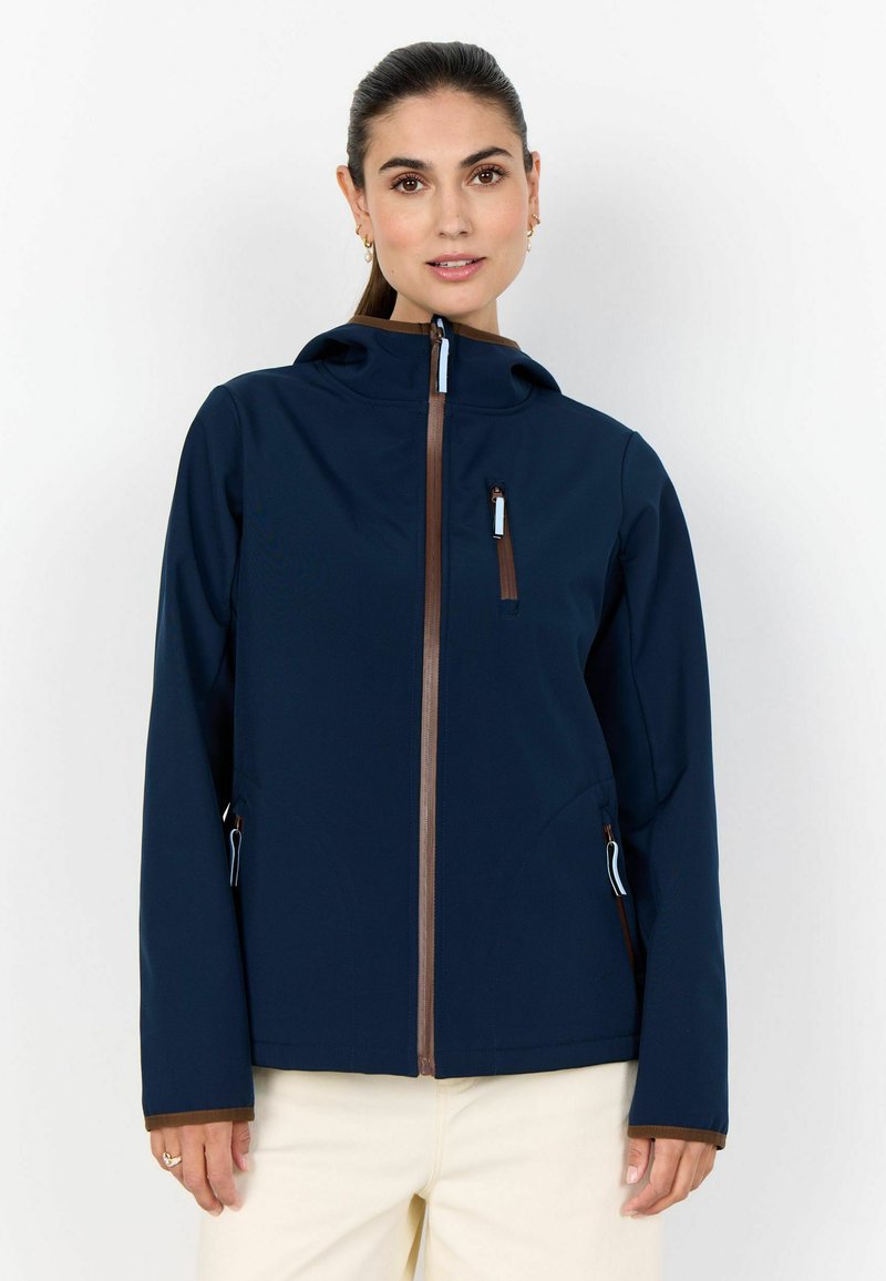 Vrouw draagt een marineblauw jack met capuchon en bruine ritssluitingen, gecombineerd met beige broek, staande tegen een effen witte achtergrond.