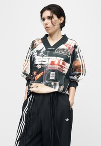 Νεαρό άτομο με κοντά σκούρα μαλλιά που φοράει πολύχρωμο αθλητικό μπλουζάκι Adidas ESPN και μαύρο παντελόνι φόρμας Adidas με λευκές ρίγες.