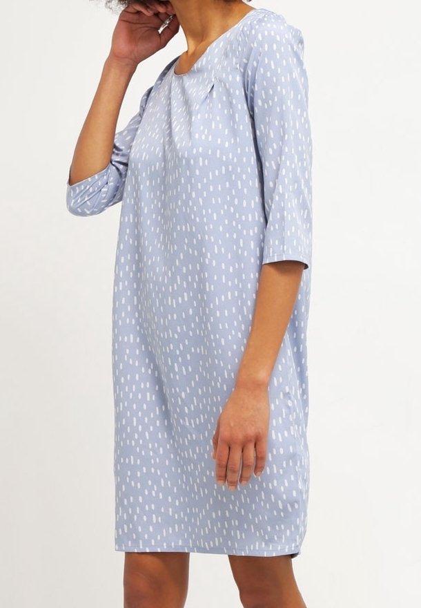 Personne portant une robe bleu clair à pois blancs, longueur genou et manches trois-quarts, debout devant un fond uni.