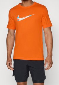 Camiseta de algodón naranja con cuello redondo. Presenta un gran logo de Nike en forma de swoosh multicolor en el pecho. Ligera, con mangas cortas.