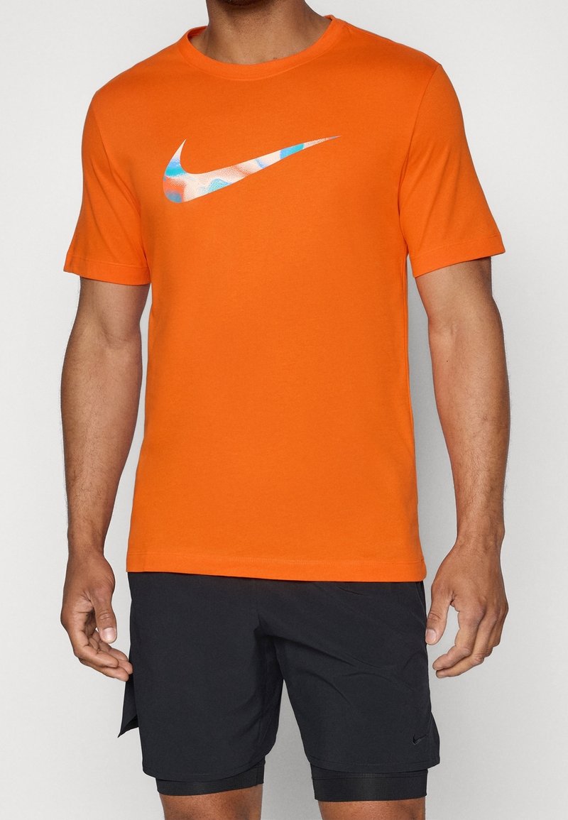 Camiseta de algodón naranja con cuello redondo. Presenta un gran logo de Nike en forma de swoosh multicolor en el pecho. Ligera, con mangas cortas.