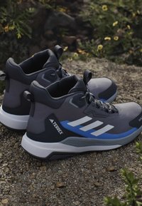 Ζευγάρι γκρι και μπλε παπούτσια πεζοπορίας Adidas Terrex με λευκές σόλες, ακουμπισμένα σε πετρώδες έδαφος με πράσινα φυτά στο φόντο.