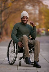 Man i rullstol iförd en grön tröja och beige byxor, svarta stövlar, grå mössa, med silvriga ringar och ansiktspiercingar, sitter utomhus.