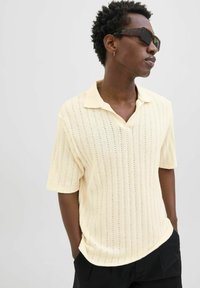 Polo shirt giallo chiaro con maniche corte, texture a righe verticali e colletto classico. Indossato con pantaloni neri, caratterizzati da una vestibilità rilassata.