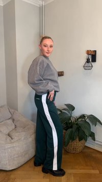 Szürke, túlméretezett pulóver és sötétzöld nadrág fehér oldalsávval. A modell egy hangulatos belső térben áll, ahol egy fonott növénytartó kosár is látható.