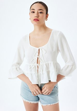 Witte blouse met lange mouwen en een vierkante halslijn, met een strikkoordsluiting en gelaagd ruffle-detail aan de zoom, gecombineerd met lichtblauwe denimshorts.