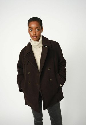 Double-breasted dark brown coat with large lapels, textured fabric, and green accent buttons, worn over a light turtleneck sweater.