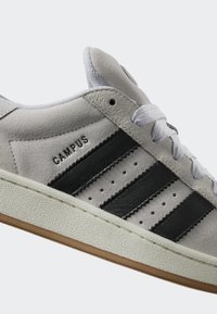 Sive dolga tenisica s tri crne pruge, teksturiranim bijelim đonom i zaobljenim vrhom. Sadrži logo s natpisom "CAMPUS" i dizajn s vezicama.