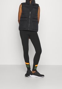 Black puffer vest with high collar and two front pockets, paired with black leggings featuring colored stripes and black athletic shoes.