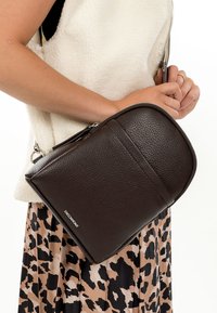 Naine, kes kannab varrukateta fliisvesti ja leopardimustrilist seelikut, hoiab tumepruunist tekstuuriga nahast õlakotti, millel on lukk.