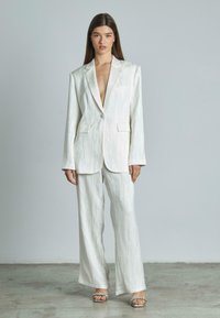 Witte textuurpak met een diepe V-hals blazer en wijde broekspijpen. Voorzien van voorzakken en een enkele knoopsluiting. Zilveren sleehakken.