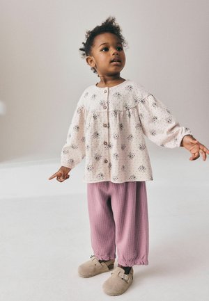 Ungt barn står med armene let strakt ud, iført en lys blomsterknappet skjorte, løse lyserøde bukser og beige slip-on sko.