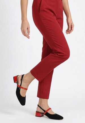 Persoon draagt rode nauwsluitende broek en zwarte slingback schoenen met lage hak, rode bandjes en blokhakken, staand op één voet.