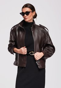 Vrouw met zwarte cat-eye zonnebril, zwarte coltrui jurk met gouden gesp riem en open ritssluiting donkerbruine leren jas tegen een witte achtergrond.