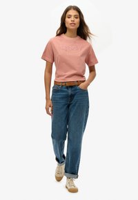 Camiseta de algodón rosa con el logo de "Superdry", combinada con pantalones vaqueros azules holgados y zapatillas beige con cordones.