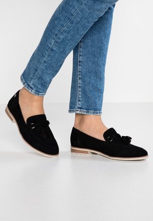 Sorte loafers i ruskind med flad sål, dekoreret med hængende kvaster og et diskret syet mønster. Brugt med blå denimjeans.