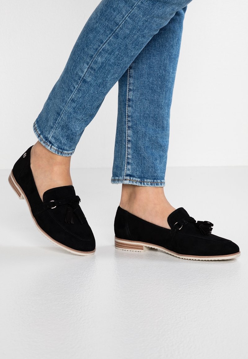 Mocassins en daim noir à semelle plate, ornés de pampilles décoratives et d'un design discrètement cousu. Portés avec un jean en denim bleu.