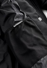Poche en tissu mesh noir sur une manche de veste avec logo blanc "SUPERDRY®" et bouton-pression métallique visible à proximité.