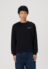 Zwarte sweatshirt van zachte stof, met een ronde hals en ribgebreide mouwen, met witte tekst "Club1990" op de voorkant.