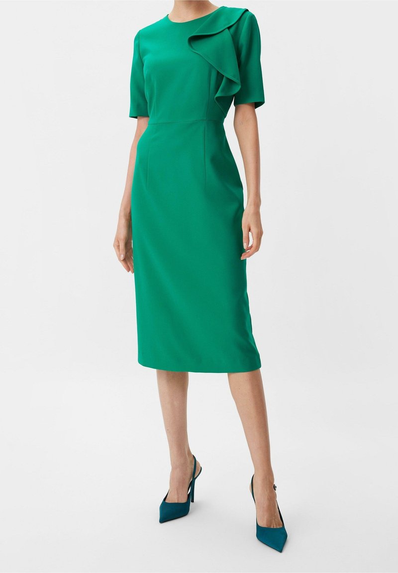 Femme portant une robe verte mi-longue à manches courtes avec un détail de volants sur une épaule, associée à des escarpins vert foncé.
