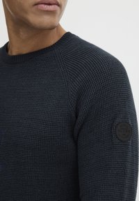 Marine blå strikket genser med teksturert vev, rund hals og en sirkulær svart logo-patch på venstre erme.