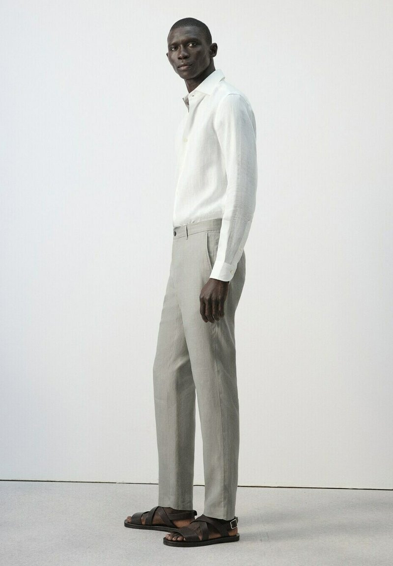 Une grande personne portant une chemise blanche à manches longues, un pantalon beige clair et des sandales en cuir marron foncé, debout devant un fond clair uni.