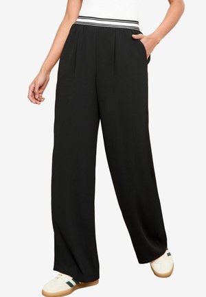 Pantaloni neri a gamba larga con una parte anteriore plissettata, caratterizzati da una vita a righe a contrasto e tasche laterali. Indossati con scarpe bianche.