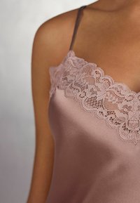 Zijdeachtige cami in zacht mauve met verstelbare bandjes, voorzien van een bloemenkantafwerking langs de halslijn voor extra detail en textuur.