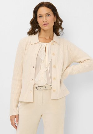 Cardigan côtelé beige clair avec un col, cinq boutons et deux poches. Porté par-dessus un chemisier en crème à volants et un pantalon assorti.