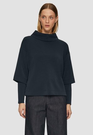 Granatowy sweter z dekoltem w cowl, szerokimi rękawami i luźnym krojem, wykonany z miękkiego materiału, noszony na koszulę z długim rękawem.