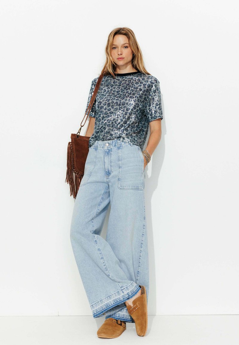 Chemise en sequins argentés avec un motif léopard, associée à un jean large bleu clair et des sabots en suède marron, accessoirisée avec un sac en franges marron.
