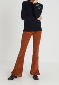 Tummansininen neulepaita pyöreällä kaula-aukolla, yhdistettynä rustinvärisiin leveälahkeisiin viitatikkihousuihin. Neulepaidan pinnassa on tekstuuria ja hienovaraisia yksityiskohtia.