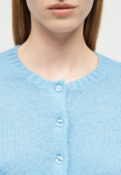 Gros plan sur une personne portant un cardigan bleu clair en tricot avec trois boutons translucides attachés, montrant le cou et le bas du visage.